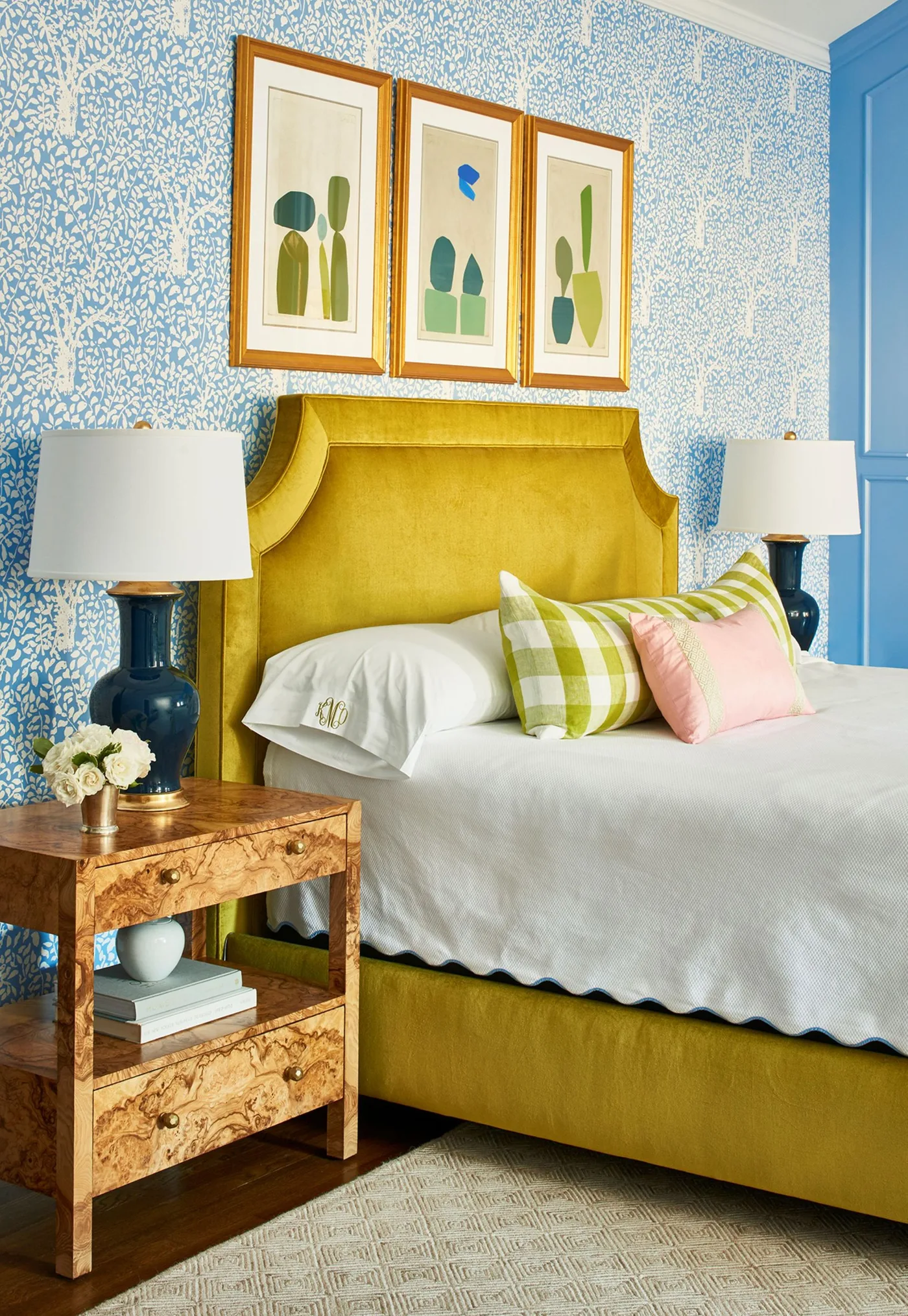 Bedroom with a shaped yellow upholstered bed, blue-and-white wallpaper, patterned cushions and a figured wooden bedside table.