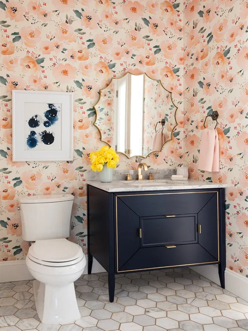 Powder room with pink-and-peach floral wallpaper, dark blue vanity, shaped metallic mirror, framed abstract picture, white toilet and pale hexagonal floor.