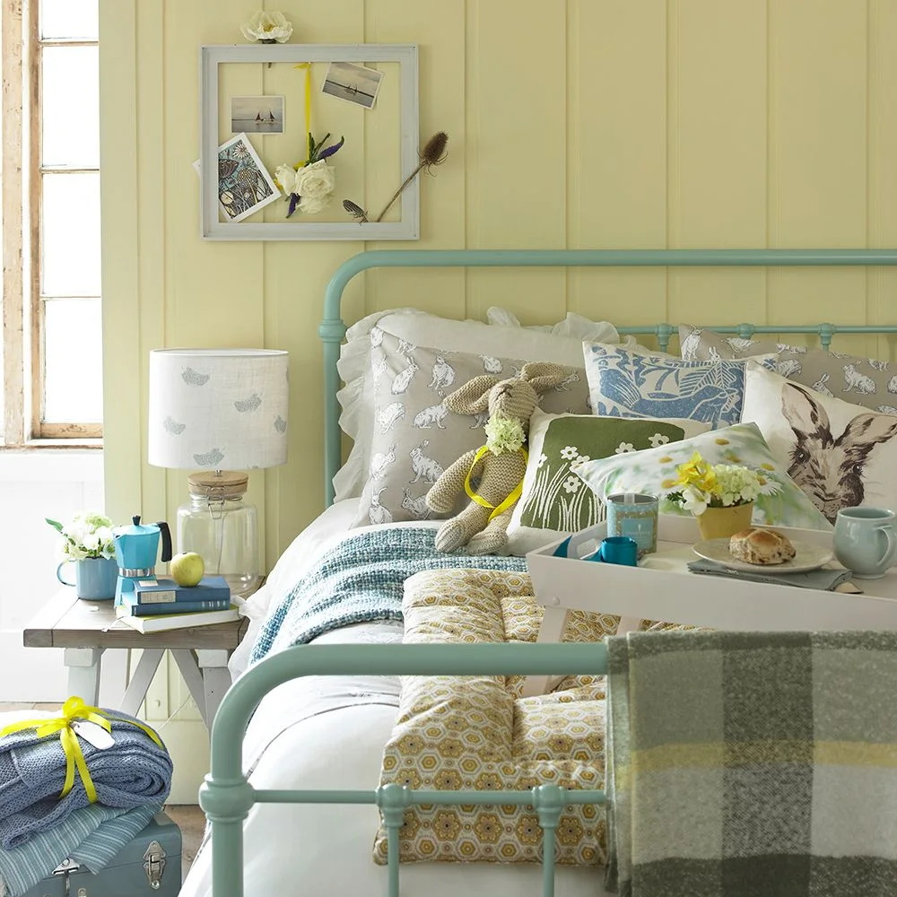 Yellow vertical paneling sits behind a mint-colored metal bed with patterned cushions, a patchwork throw and a white breakfast tray.