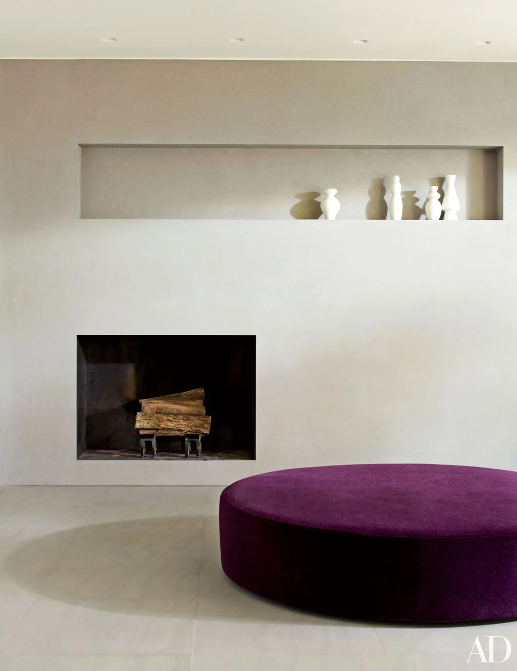 A dark rectangular fireplace opening sits within a plain pale wall below a long recessed shelf holding white vessels beside a rounded purple ottoman
