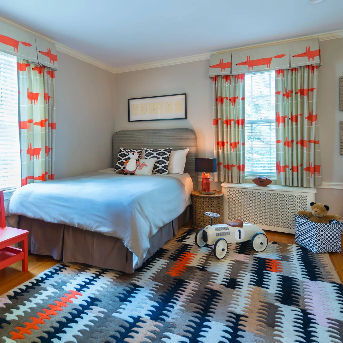 A blue, gray, orange and white patterned rug sits beneath a single bed beside curtains decorated with orange foxes.