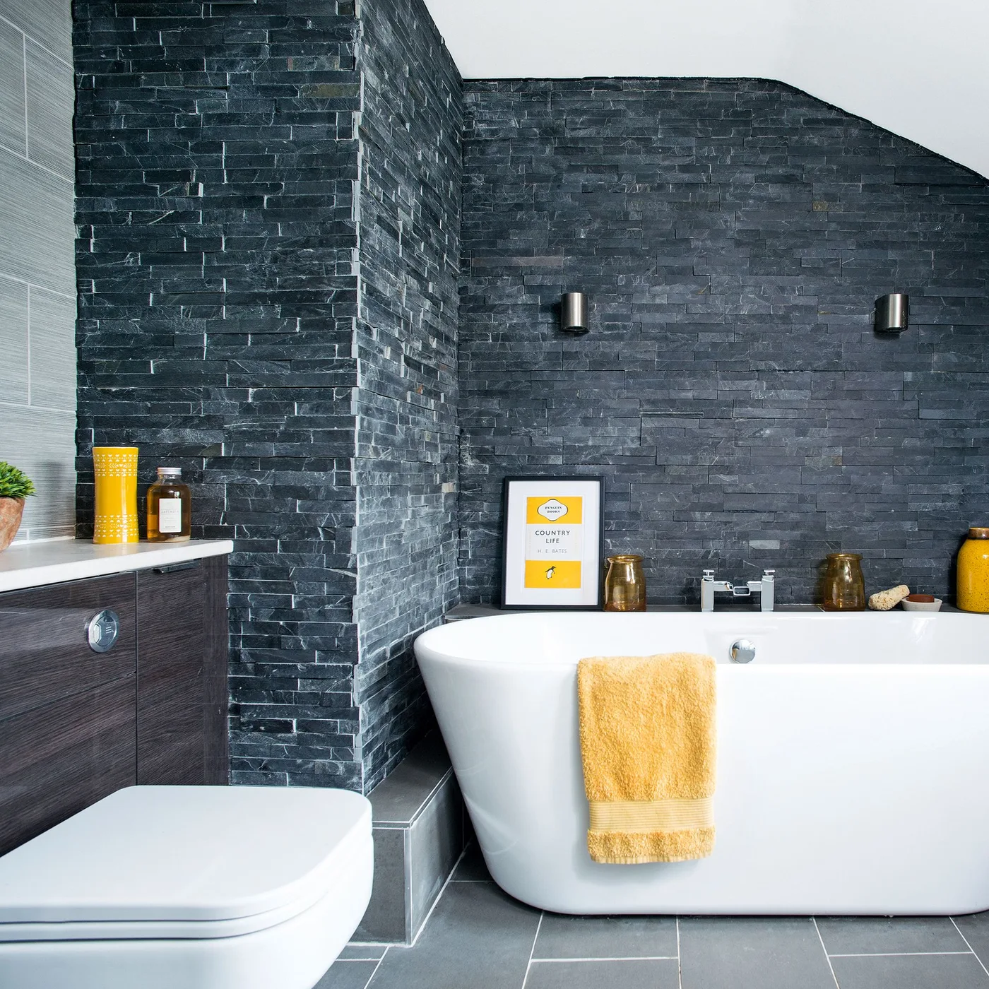 Bathroom with dark gray textured small wall pieces, white oval bath, gray floor, mustard towel and accessories, wall lights, and pale ledge.