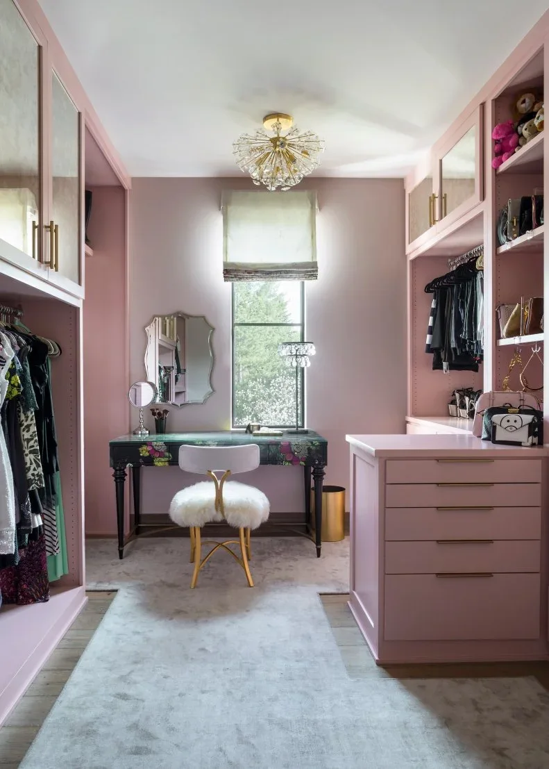 Pink closet cabinetry frames open hanging rails and drawers beside a small dressing table under a window.
