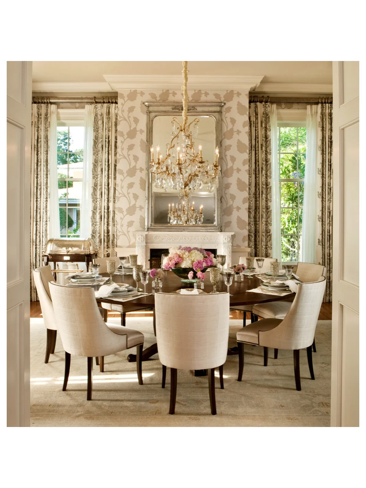 A restrained patterned background frames a round dining table, pale chairs and a substantial chandelier.