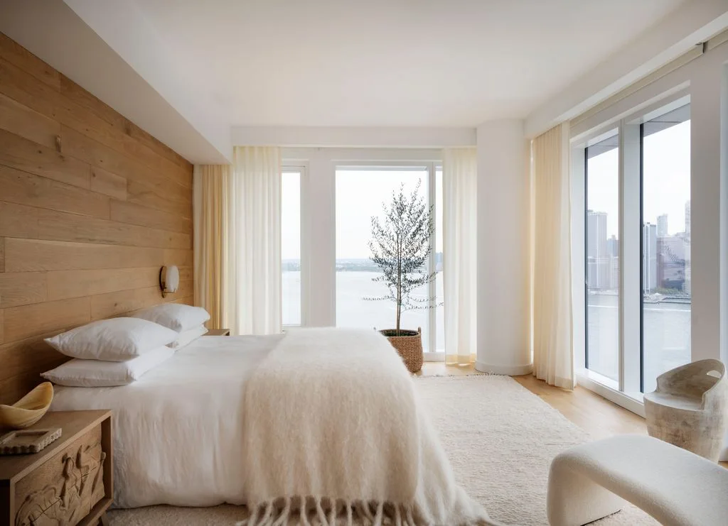 A pale bed with white bedding stands against horizontal wood wall boards beside broad glazed doors, light curtains, a pale rug and a small chair