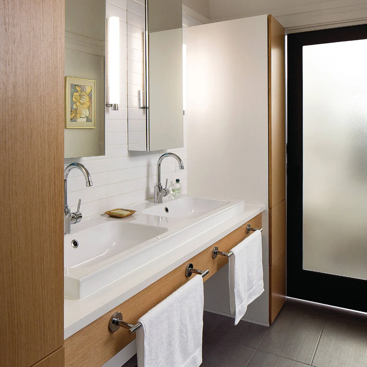 Two short chrome rails hold white towels on the wood front beneath a paired white basin arrangement and tall faucets.