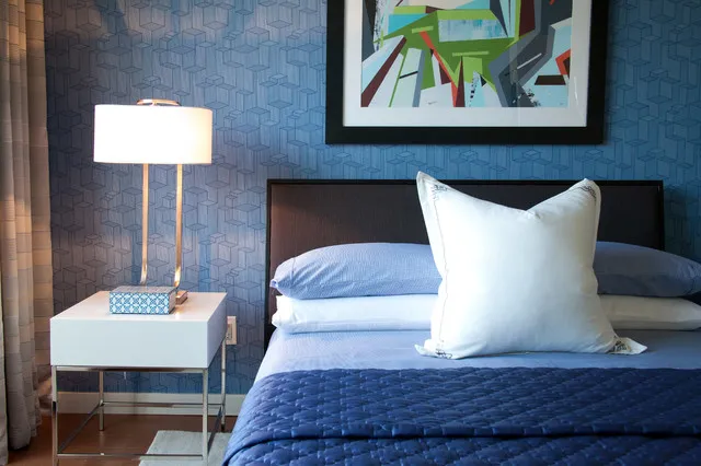 Blue geometric wallpaper frames a dark headboard with blue-and-white bedding, a white table and a slender brass-colored lamp.