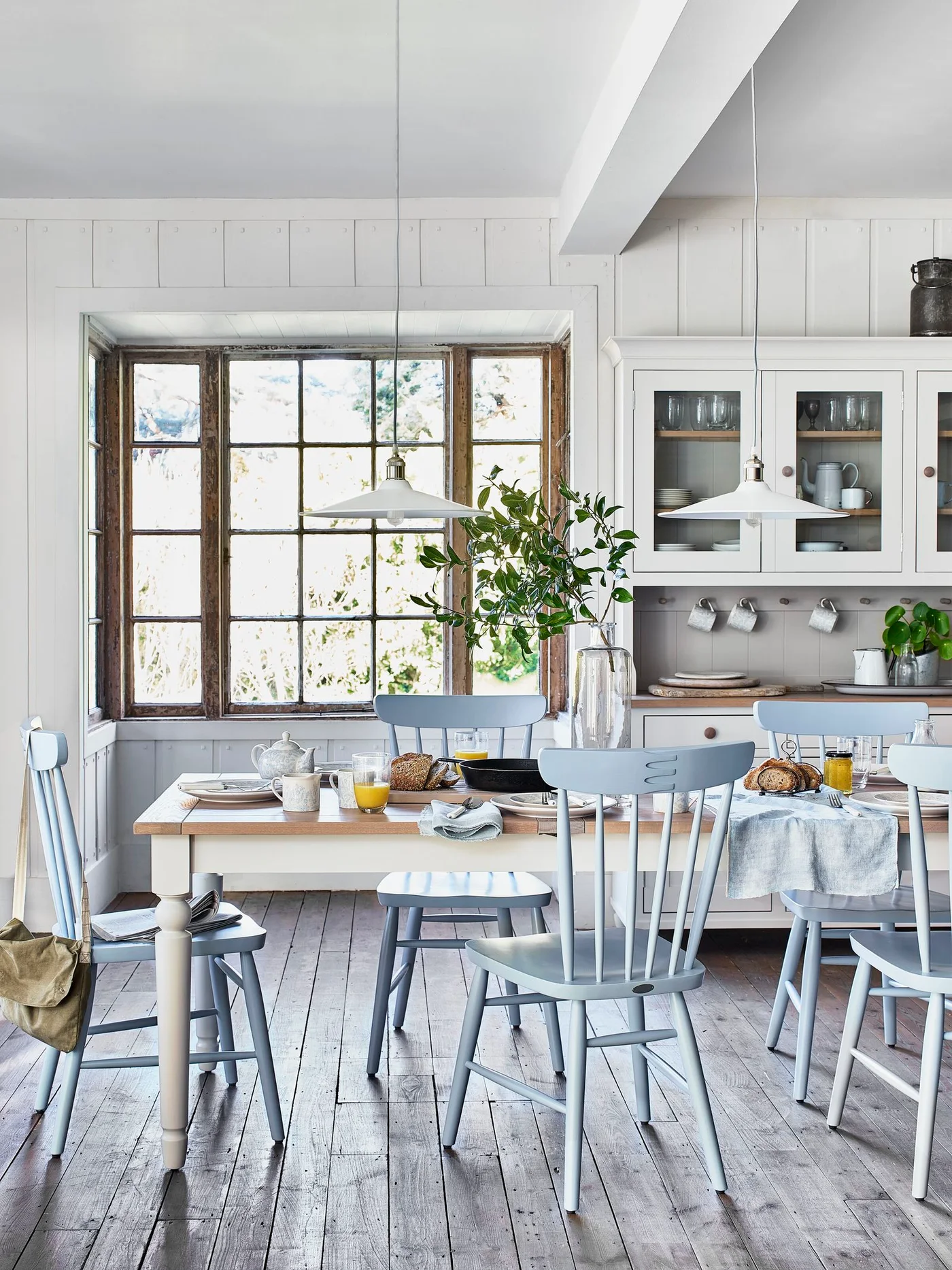 Pale cream kitchen storage and a cream-painted dining table with blue chairs, glazed upper cupboards and an old wooden window frame.