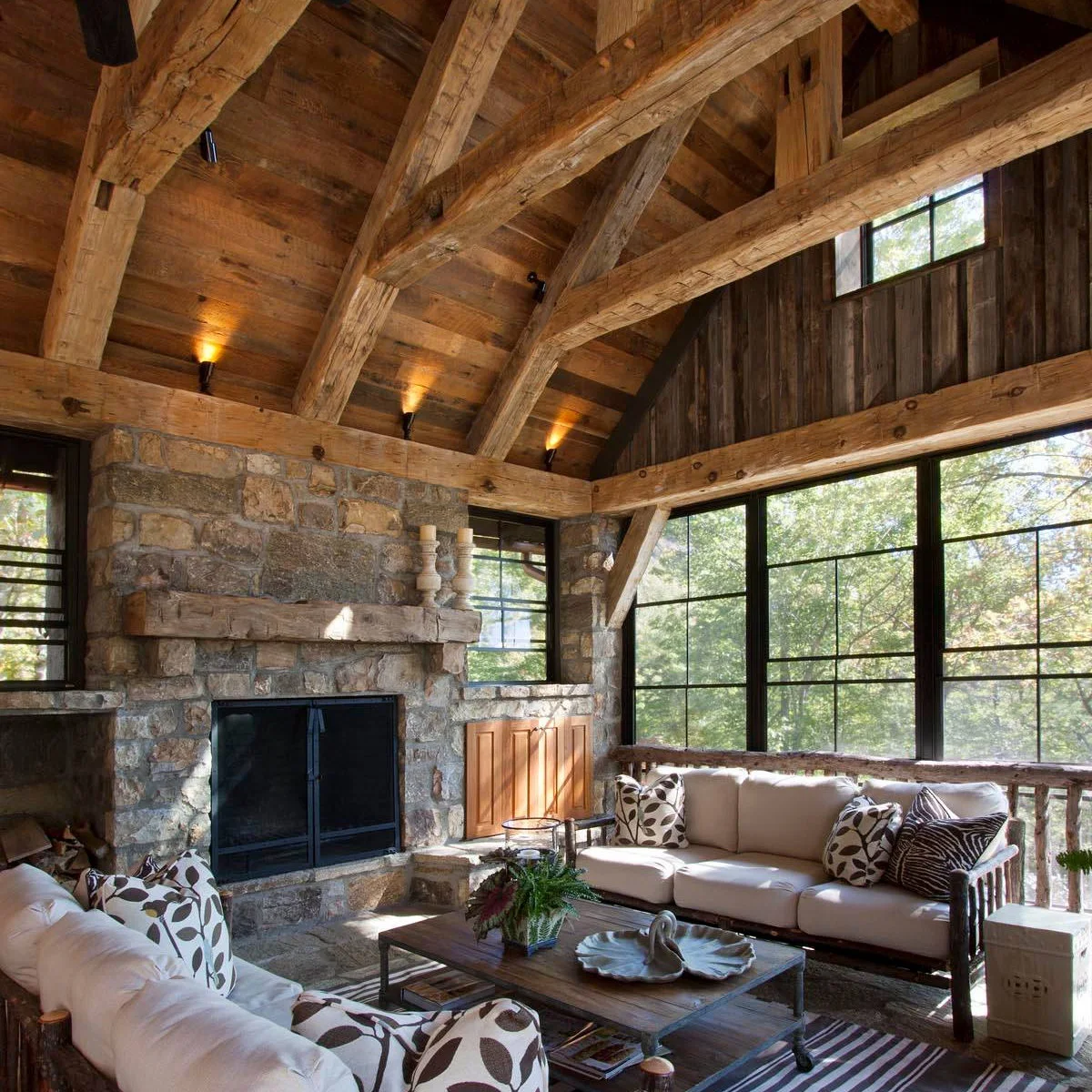 A rustic enclosed porch with exposed wood roof framing, stone fireplace, pale sofas and black-framed windows.