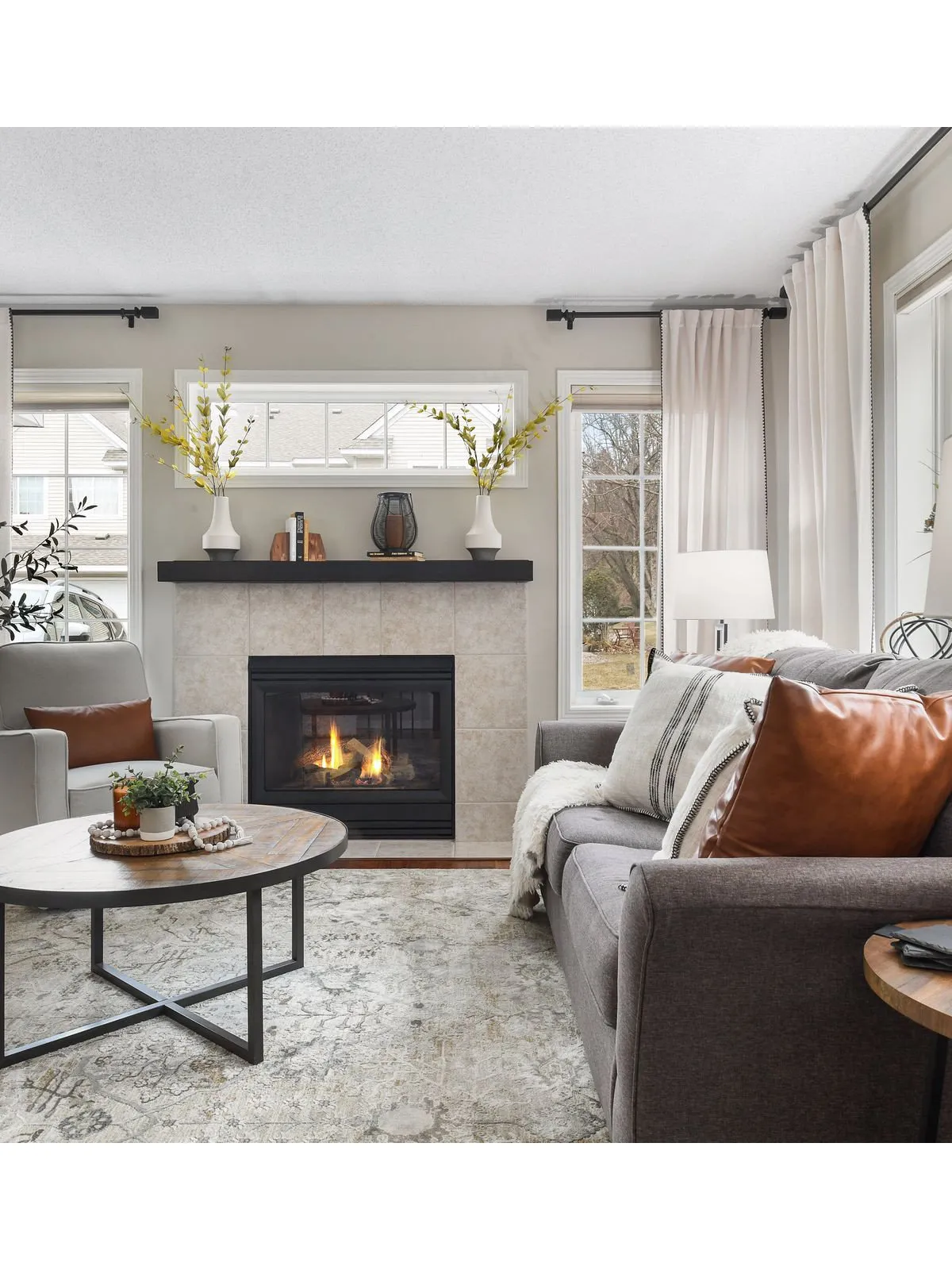 A gray sofa with white and rust-colored cushions sits beside a bright fireplace and pale curtains.