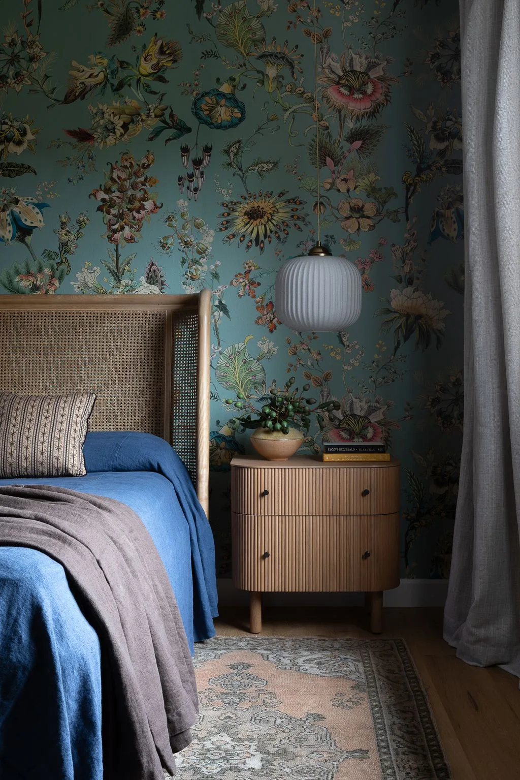 Deep green floral bedroom wallpaper with a cane headboard, blue bedding, gray throw, ribbed wooden bedside cabinet, and white hanging light.
