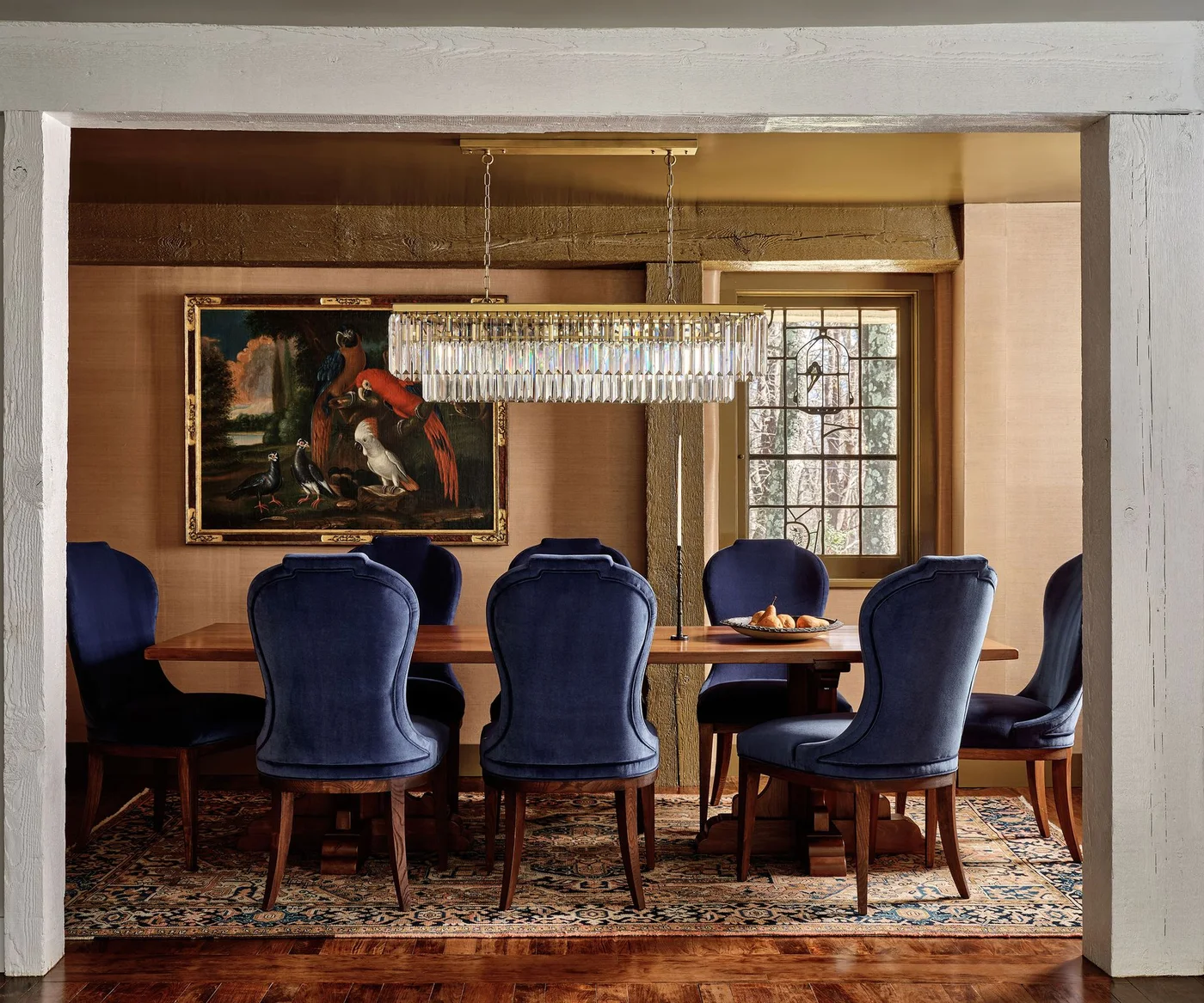 Deep blue upholstered dining chairs surround a wooden table beneath a long clear hanging light beside framed art and warm walls