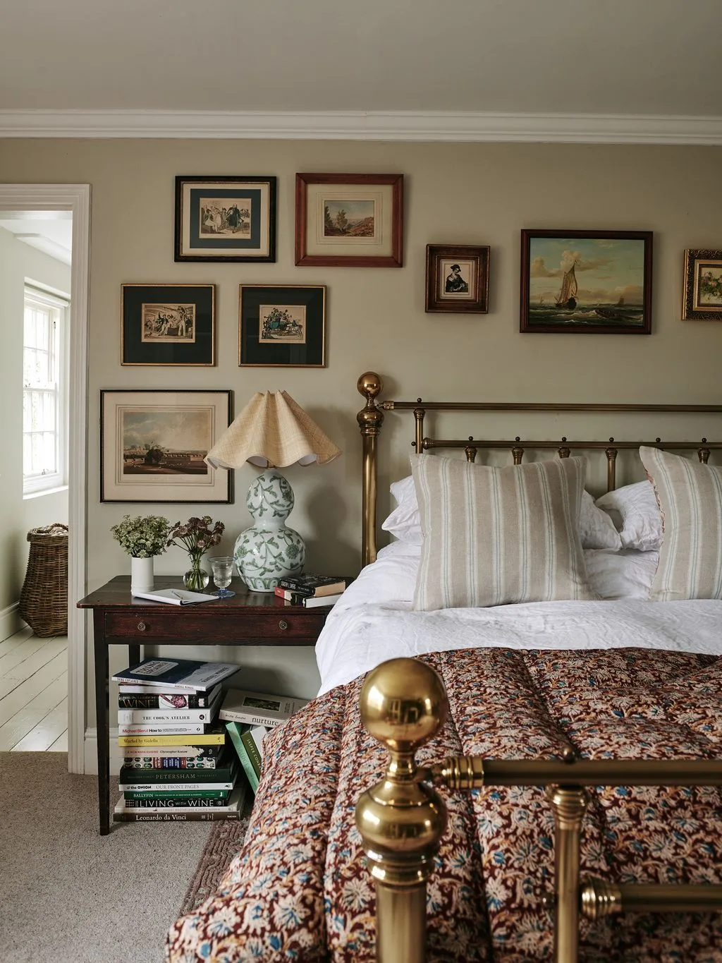 Bedroom with gold-colored metal bed, striped pale cushions, patterned bed cover, framed picture group and green-patterned lamp