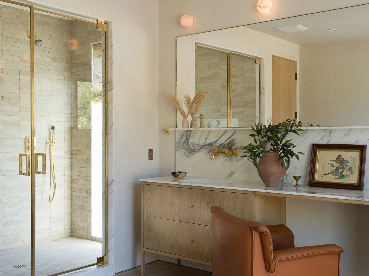 A tan chair sits beside a timber vanity and pale veined backsplash near a glass shower.