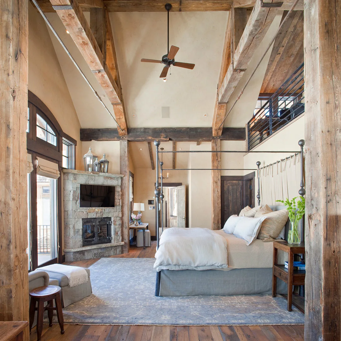 A dark metal four poster bed beneath exposed beams beside a stone fireplace and pale rug