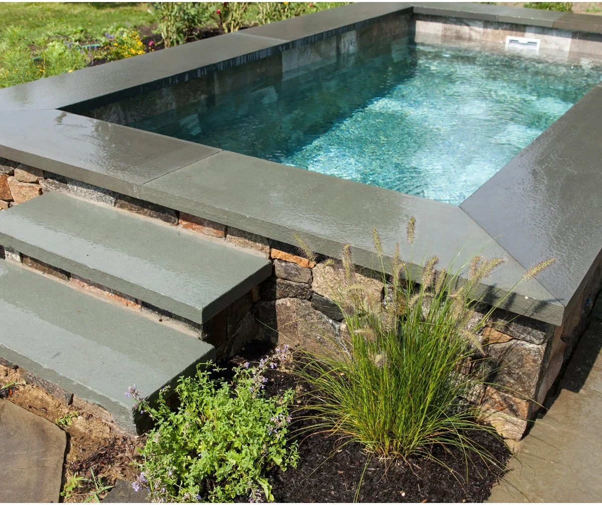 A small raised rectangular pool with a stone-clad outer face and gray coping sits among grasses and garden planting.