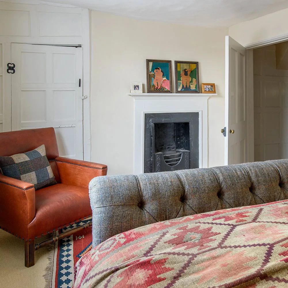 Bedroom with a gray upholstered bed, red patterned bedcover, orange armchair, pale walls and framed pictures on a fireplace mantel.