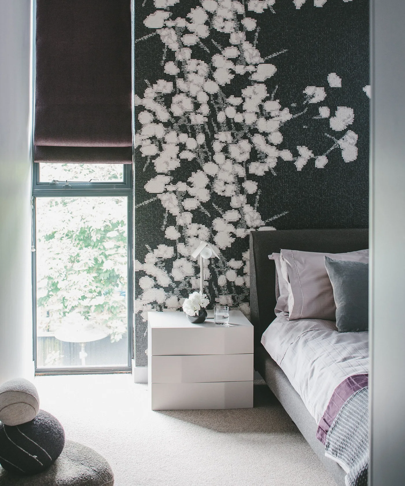 White flowering branches on a dark mural beside a tall window and a pale bedside chest.