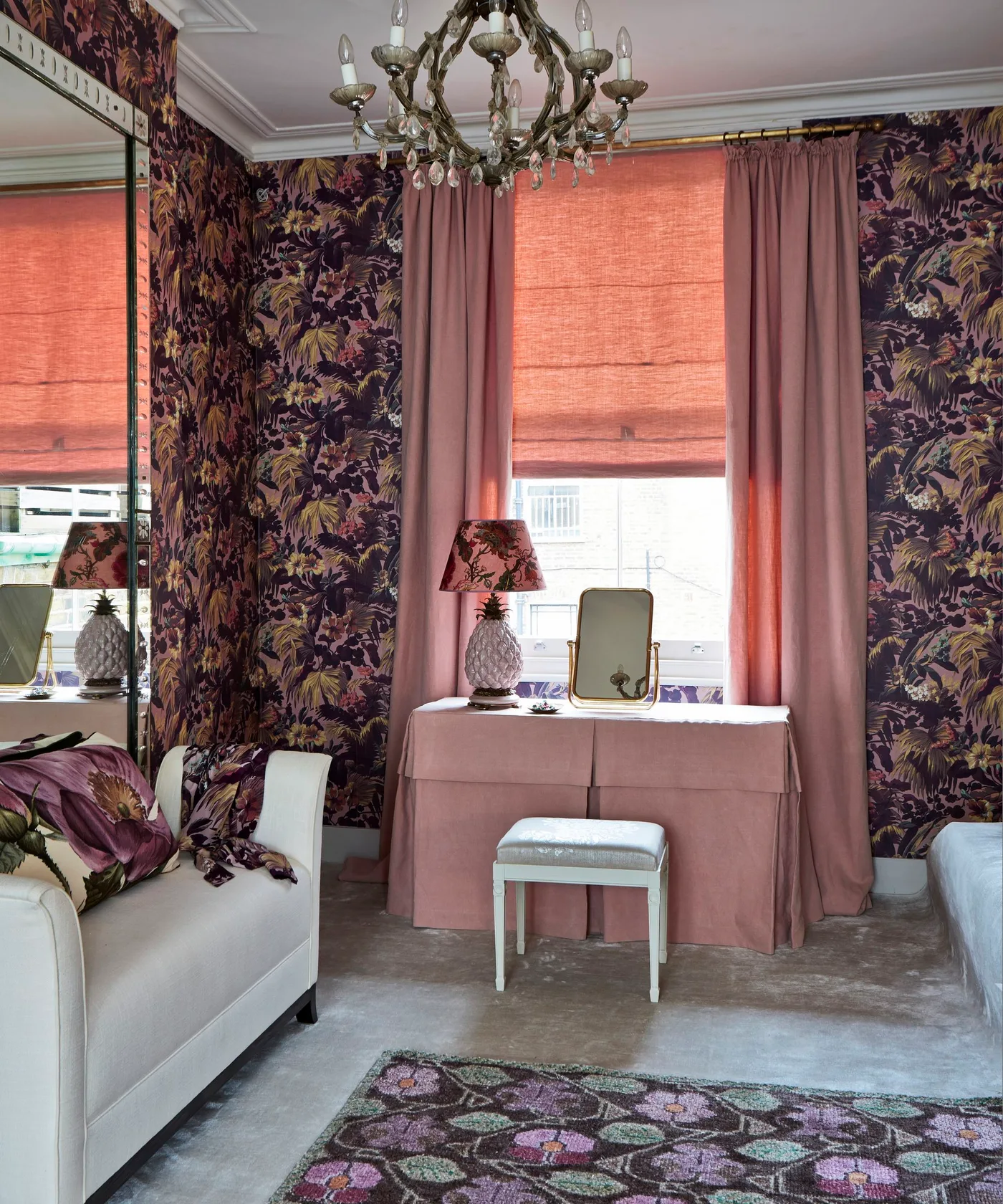 Bedroom with dark floral walls, pink dressing table, pale stool, pink curtains and blind, large mirror and ornate chandelier.