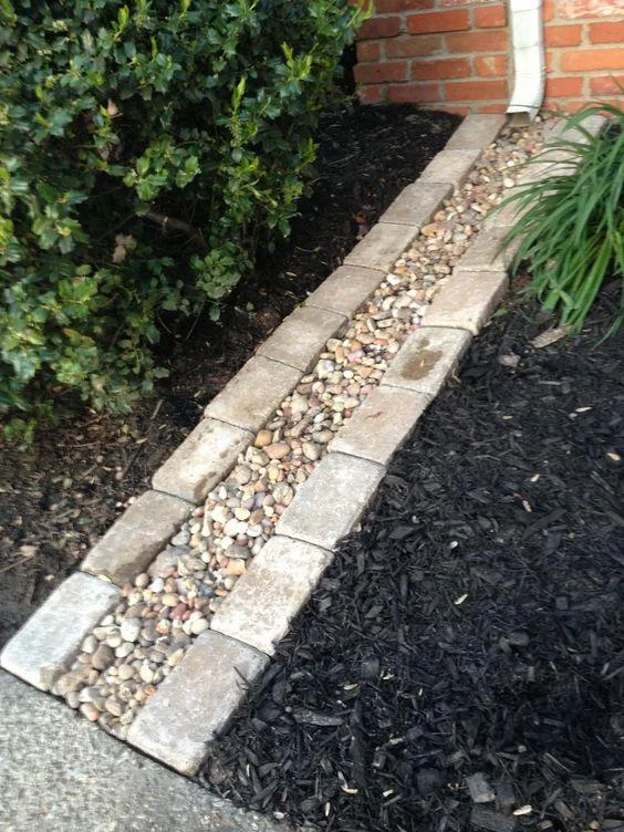 A narrow stone-filled channel edged with rectangular blocks runs from a downspout through dark mulch