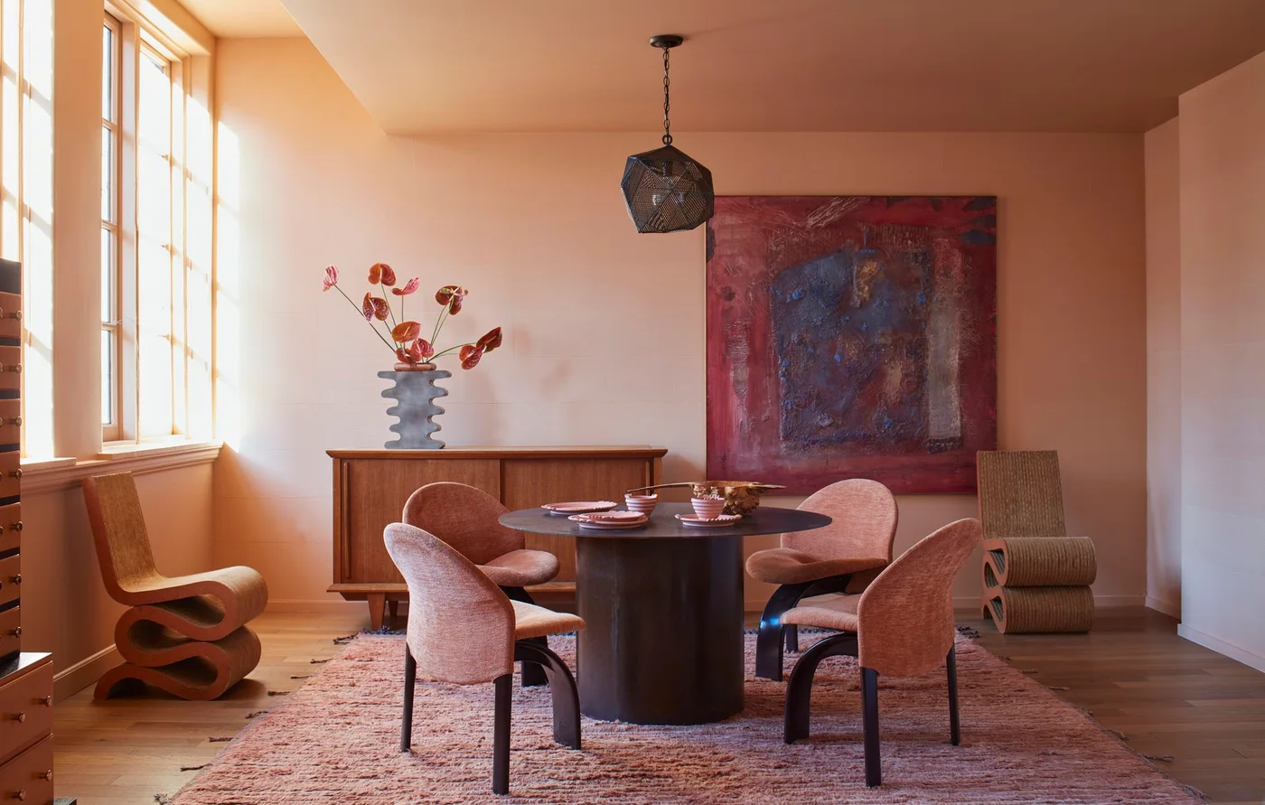 A peach dining room has a dark round pedestal table, rounded pink chairs, a pale wood sideboard, a dark pendant and a large red abstract artwork