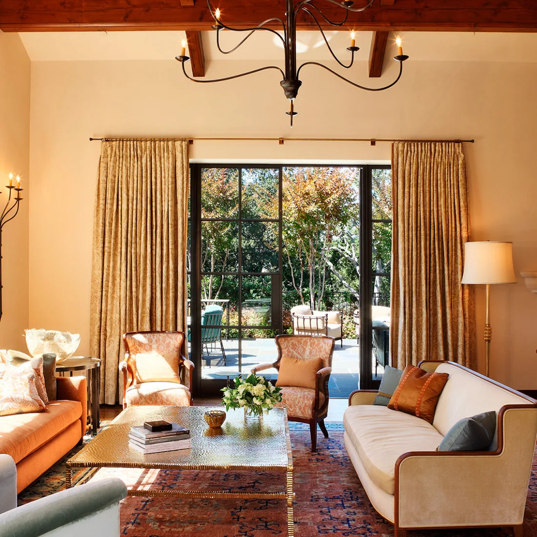 Warm orange and pale living-room seating surrounds a low reflective table beneath wooden beams and a dark curved chandelier beside tall glazed doors