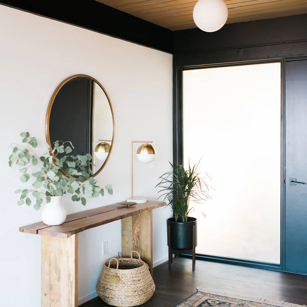 A narrow wood console beneath an oval mirror, with a small lamp, white vase and woven basket below.