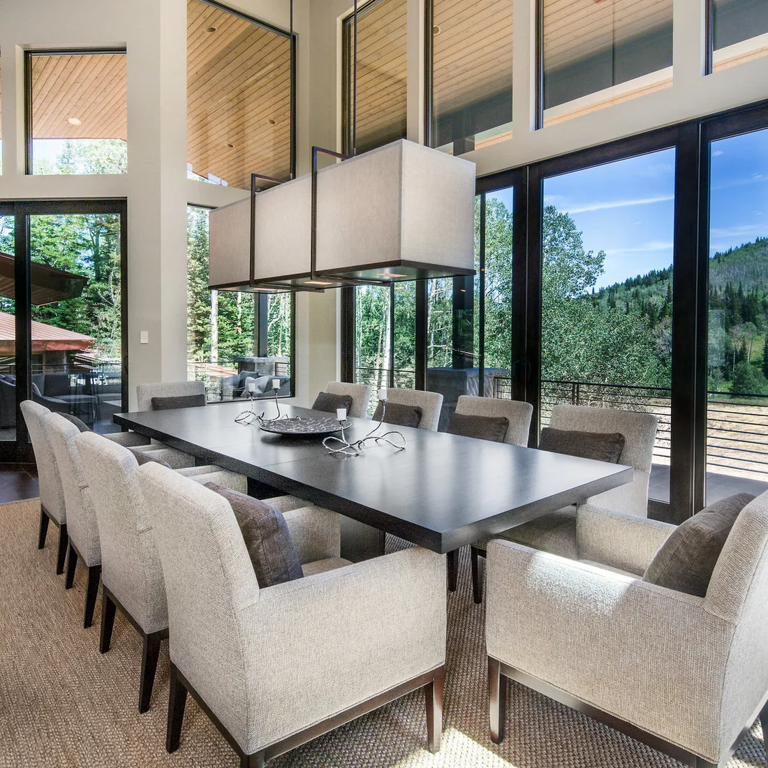 Neutral textured rug under a dark dining table and pale upholstered chairs beside tall black-framed windows