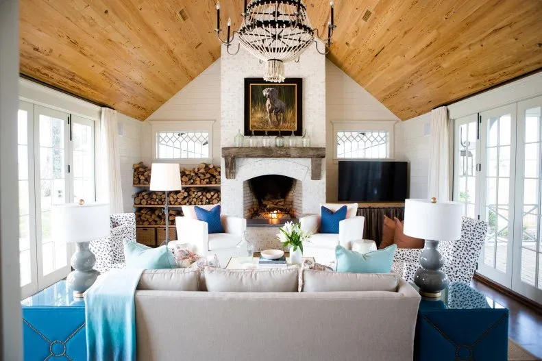 A pale brick fireplace rises under a timber vaulted ceiling with log shelves on one side and a television on the other.