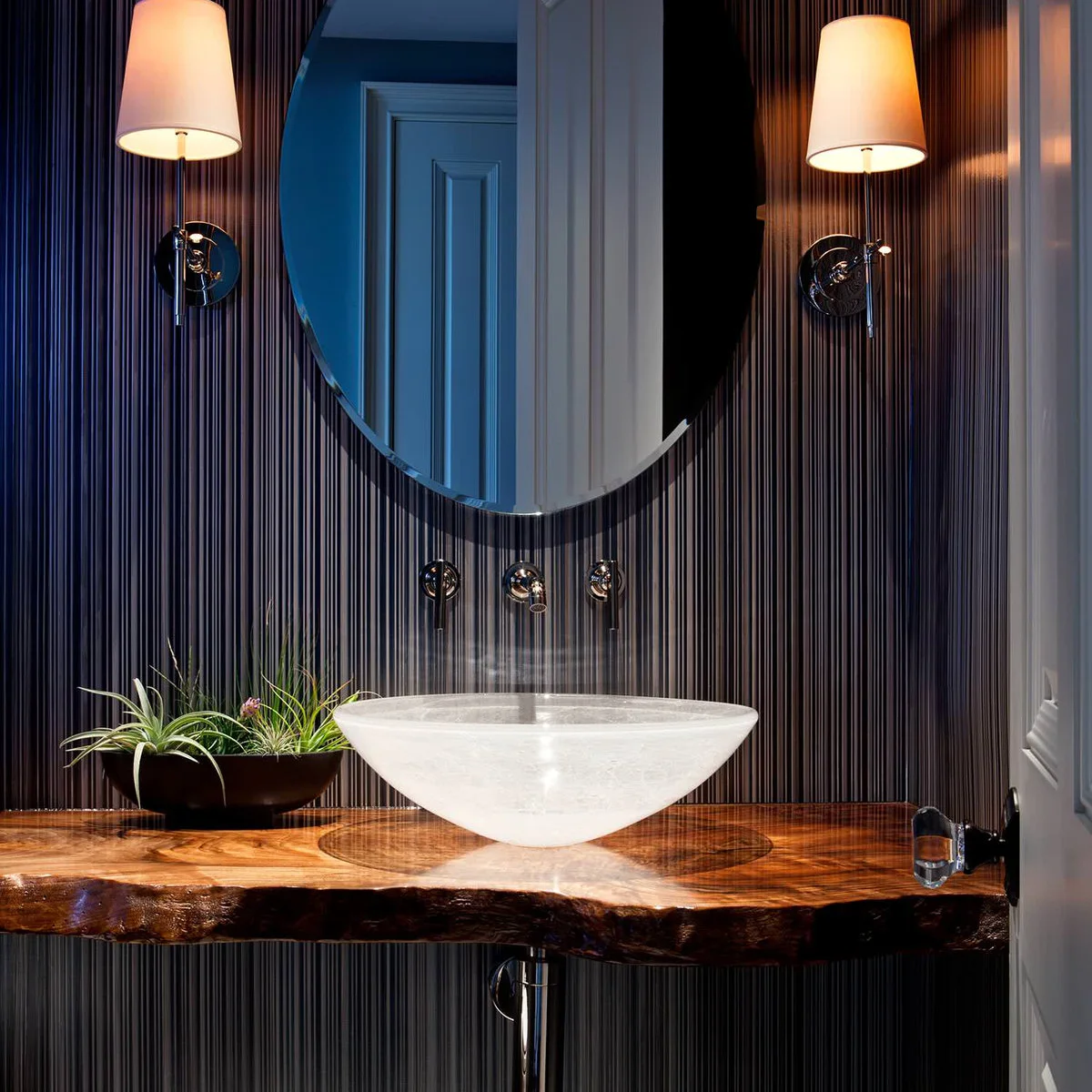 A pale translucent vessel bowl sits on an irregular-edged wood counter beneath an oval mirror and shaded wall lights against striped walls.