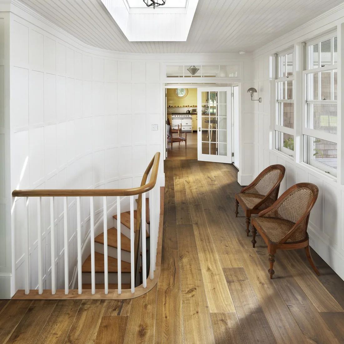 White wall paneling and a white stair rail surround a wood-floored landing with two cane chairs below windows and a rooflight.