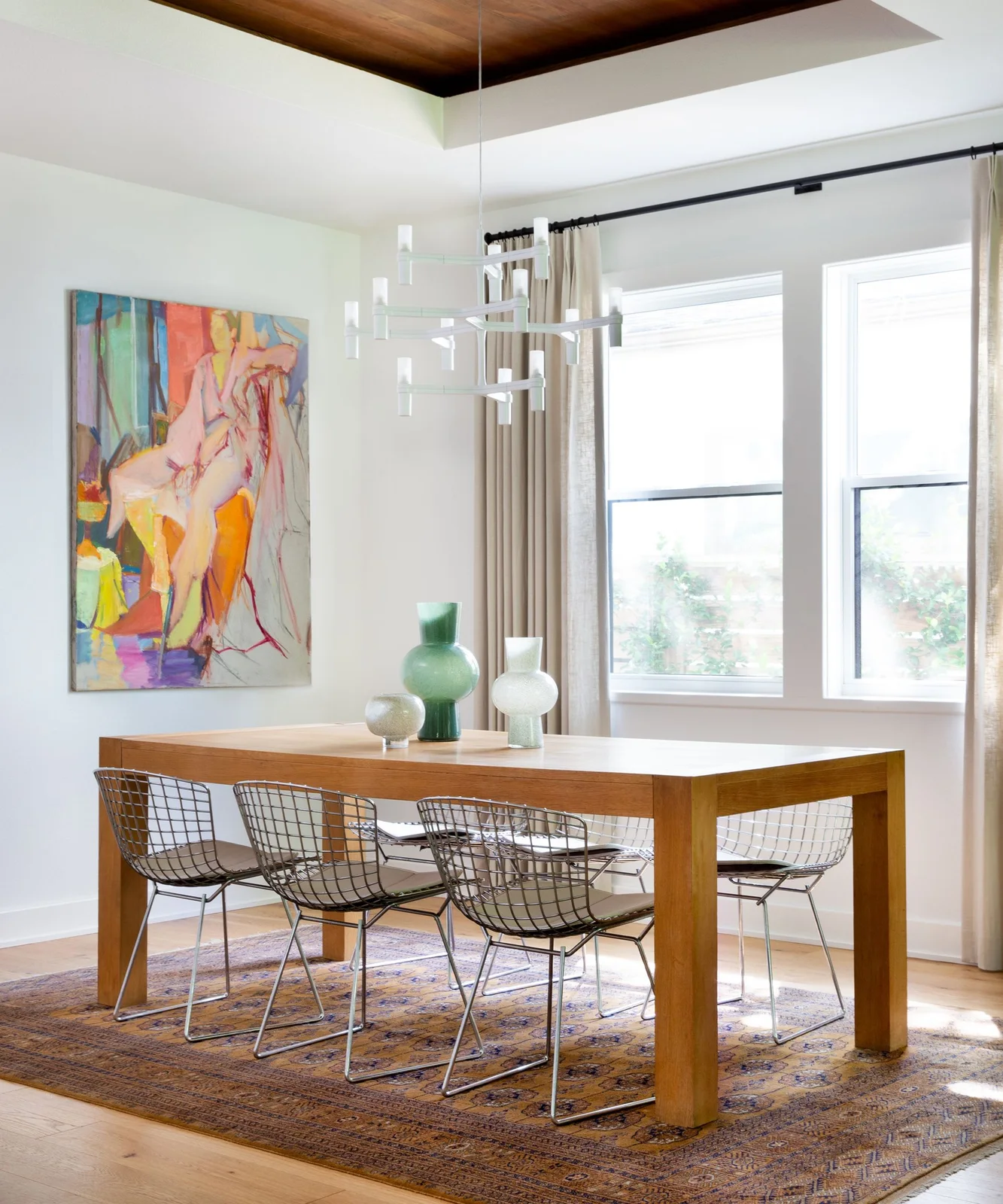 A wooden dining table and wire-frame chairs sit beneath a white branching light beside large colorful artwork and pale curtains