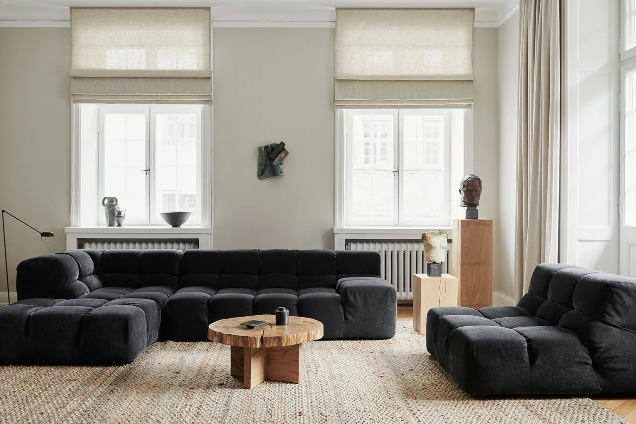 Dark gray modular sofas surround small rounded wooden tables in a light room