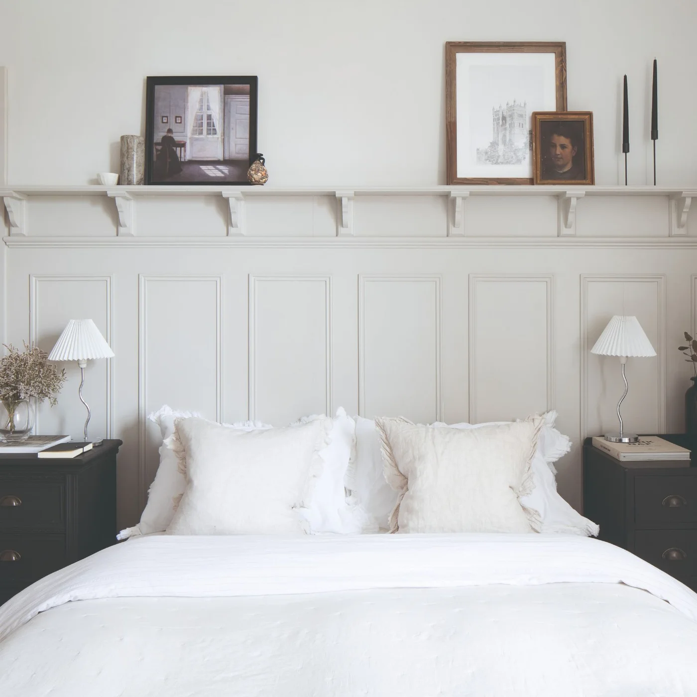 A white bed sits between dark drawer tables and matching pale lamps beneath tall white paneling and a narrow display ledge with framed pictures.