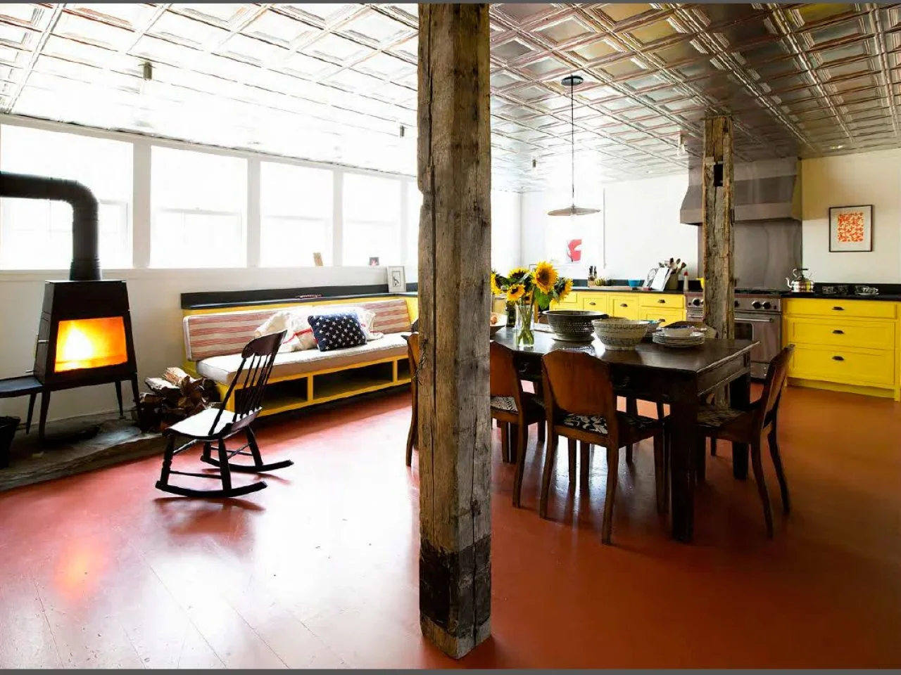 Yellow kitchen cabinets and a dark dining table sit between rough timber posts beneath a patterned pale ceiling.