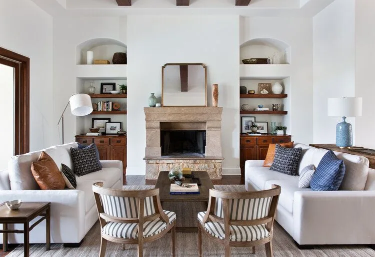 Pale stone fireplace between white sofas, arched shelf recesses, wooden cupboards and striped chair backs.