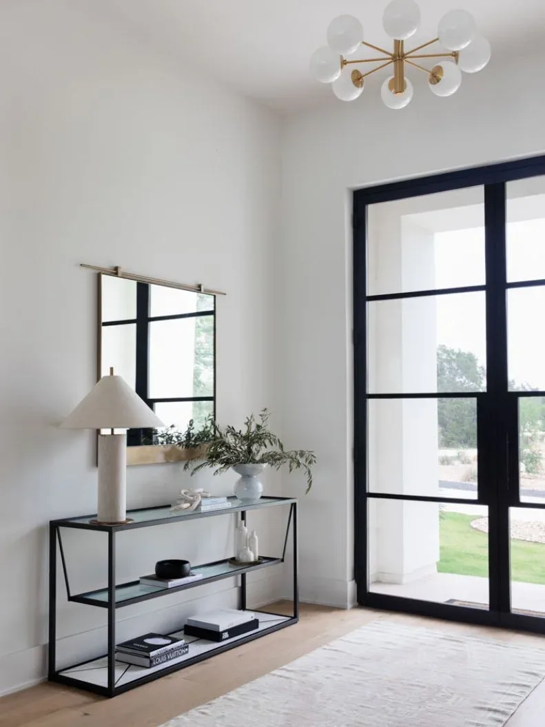 Gold-colored branching globe chandelier above black-framed glazed entrance, slim black console, pale lamp and rectangular mirror