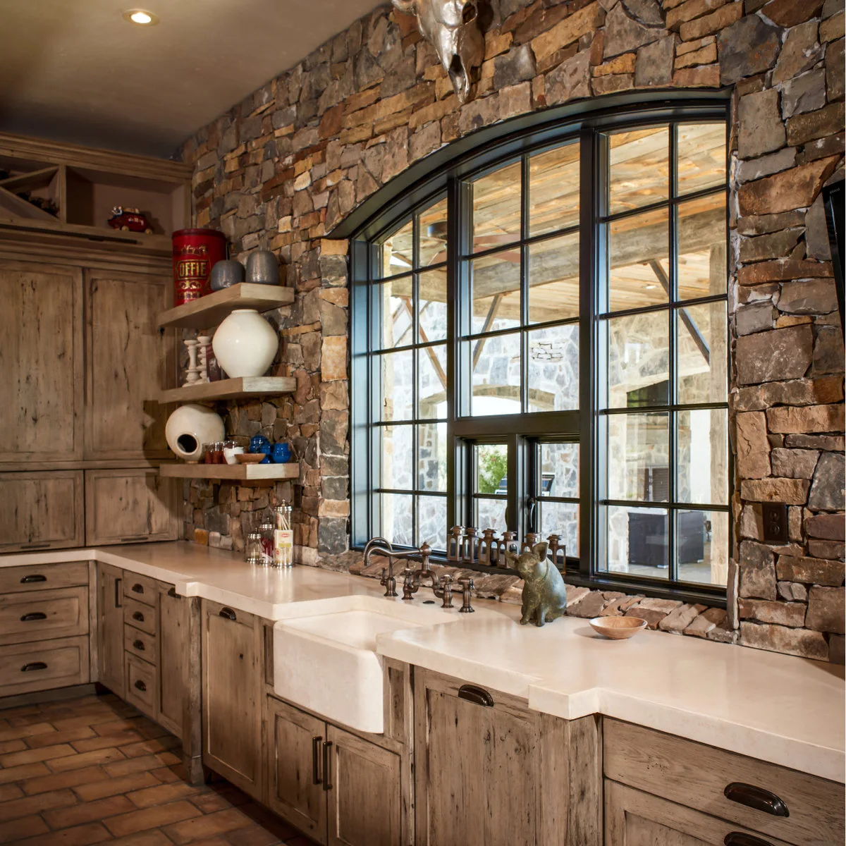 A dark arched kitchen window set in a stone wall above a pale apron-front sink and rustic wood cabinets.