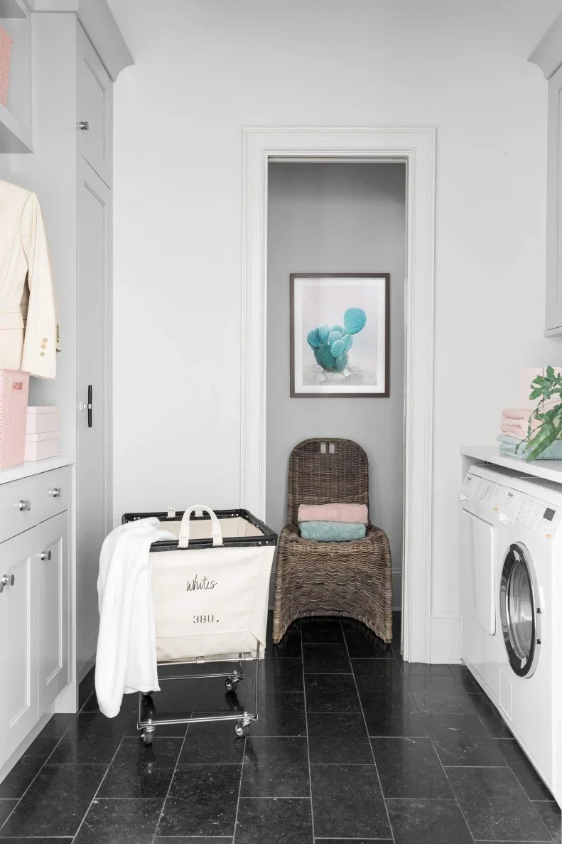 A gray laundry room has side-by-side appliances, fitted cupboards, a wheeled fabric hamper and a pocket doorway leading to a small adjoining room.
