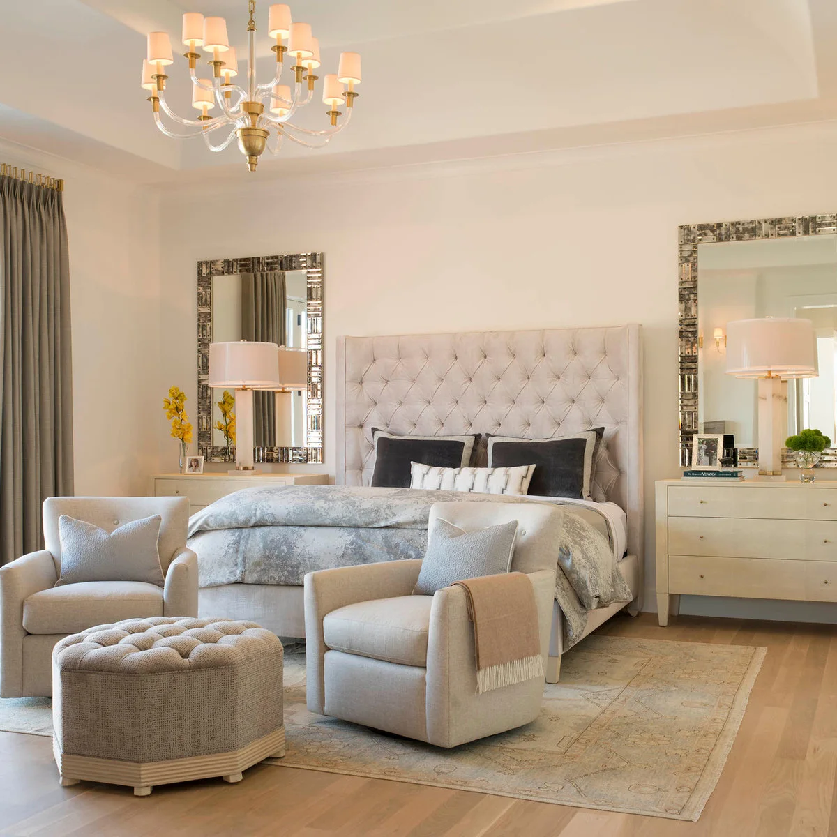 Two pale armchairs and a tufted ottoman sit at the foot of a bed with matching bedside chests and mirrors.