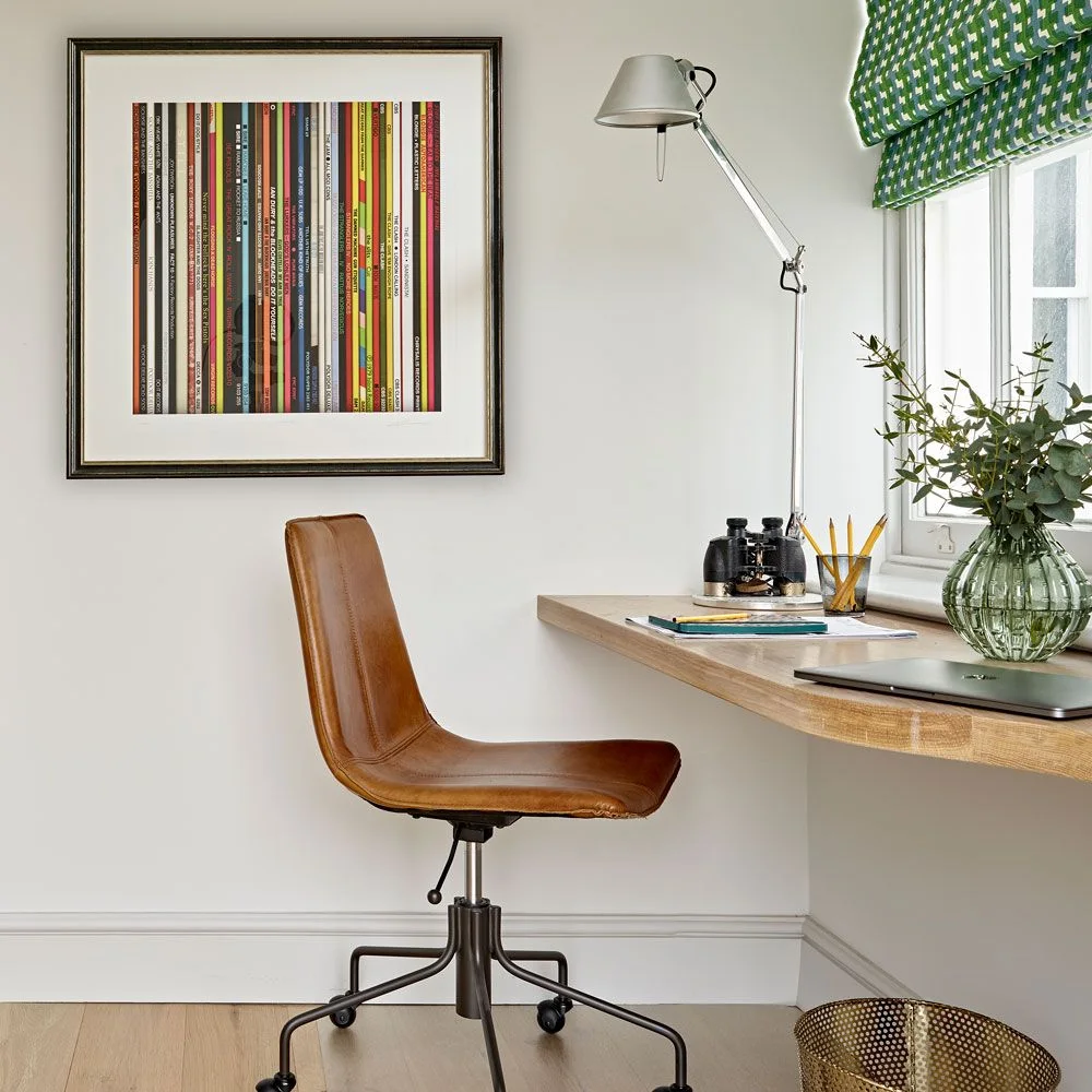 A thick wood worktop with a rounded corner sits beside a white wall and window, with a brown swivel chair, an articulated metal lamp and a framed striped print.