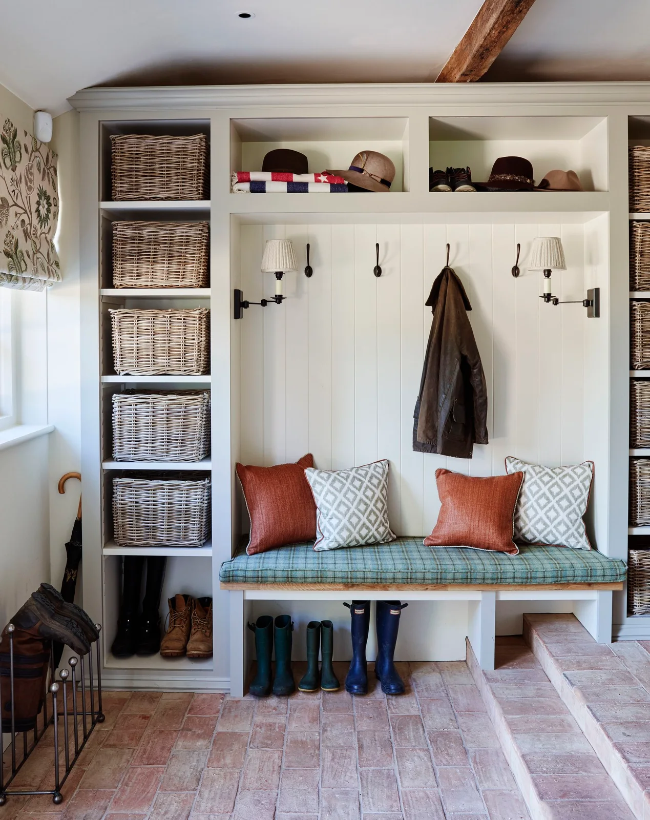 Pale vertical boards and coat hooks surround a cushioned wood bench beside tall basket shelves, upper open compartments and a visible ceiling beam