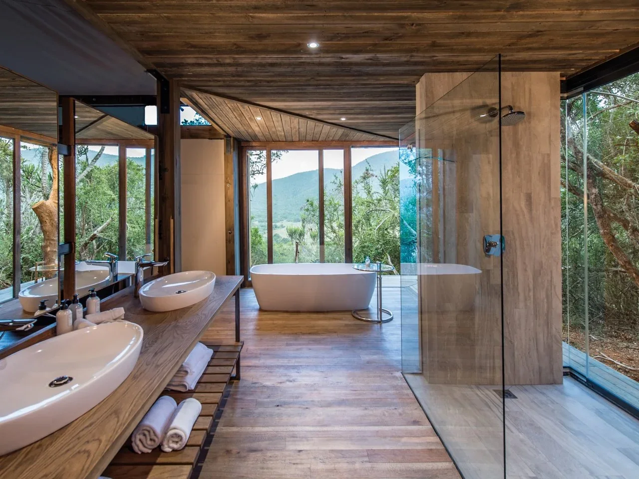 Timber surfaces and large windows surround a white tub beside a glass shower with a wood-toned tiled floor.