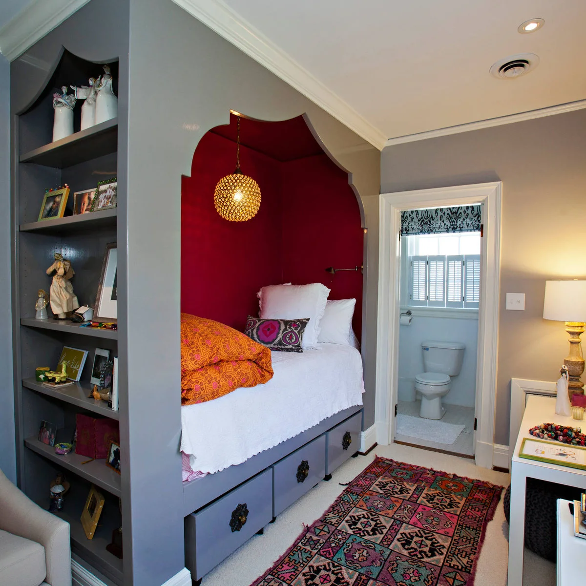 Gray built-in bed alcove with deep red interior, orange textiles and drawers below