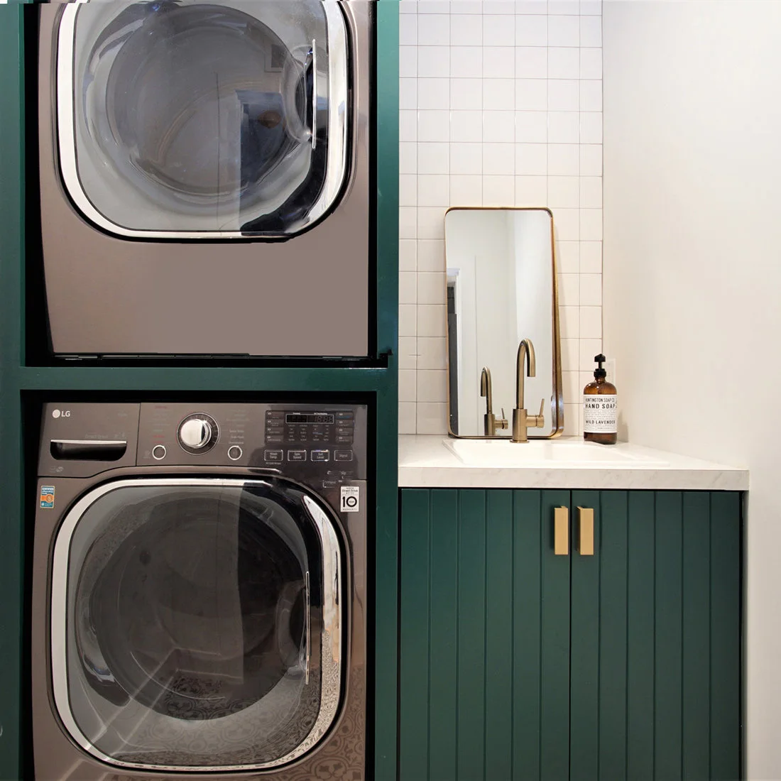 Deep green cabinets surround stacked gray machines beside a white counter, sink, warm-metal tap and rectangular mirror.
