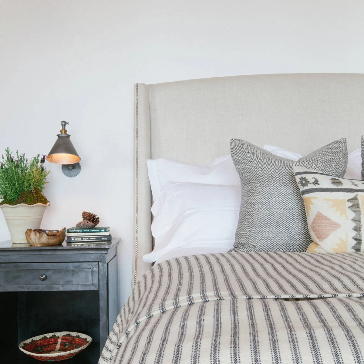 Pale upholstered headboard beside a wall reading light and grey nightstand with an open lower shelf