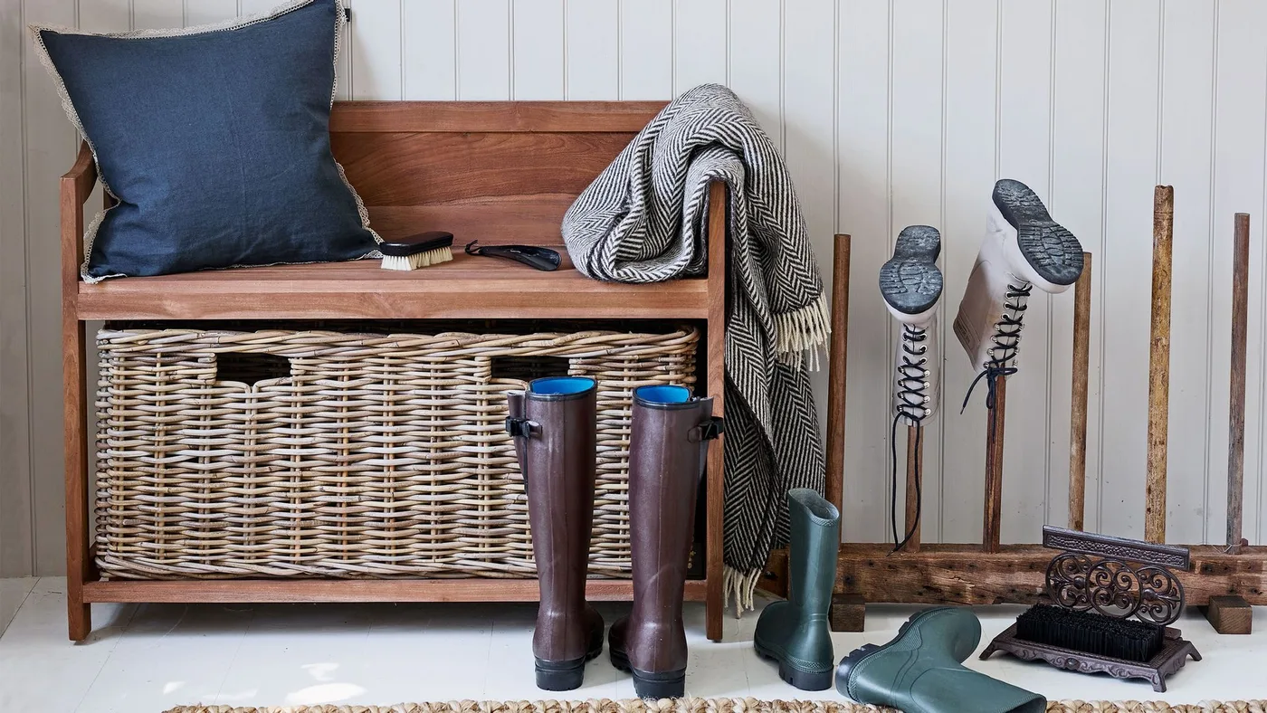 Wooden entryway bench with large woven storage basket, navy cushion, striped throw and boots against white paneling.