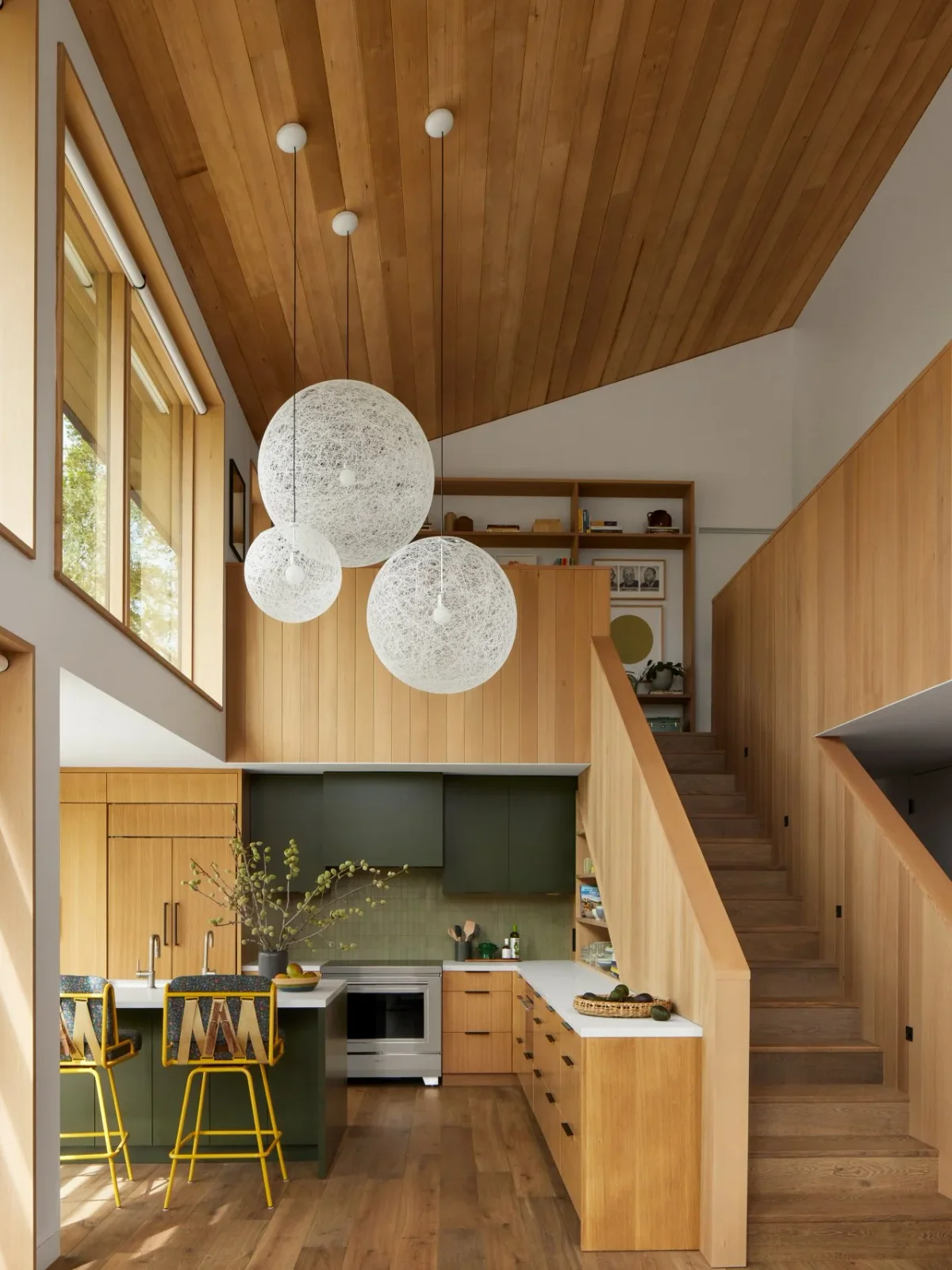 Warm white oak cabinetry sits beneath a sloping timber ceiling, with a dark green hood, pale counters and yellow-framed stools.
