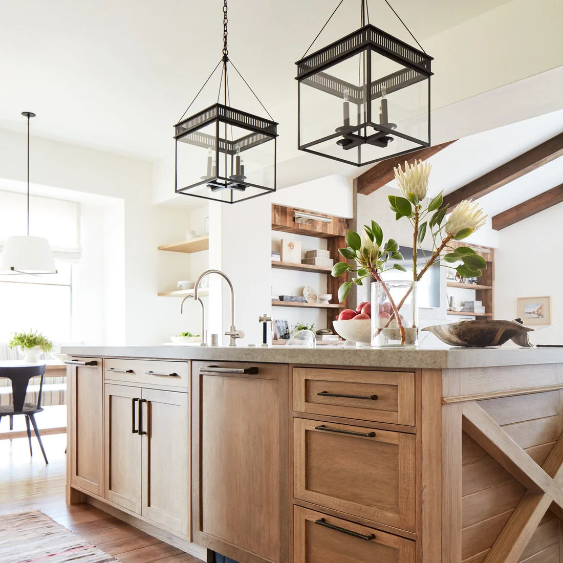 A warm timber island with dark handles sits below black lantern pendants beside timber shelving and ceiling beams.