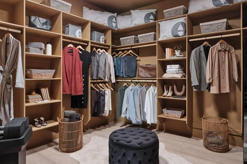 A timber walk-in closet has double hanging rails, open shelves, top storage bags and a dark round ottoman.